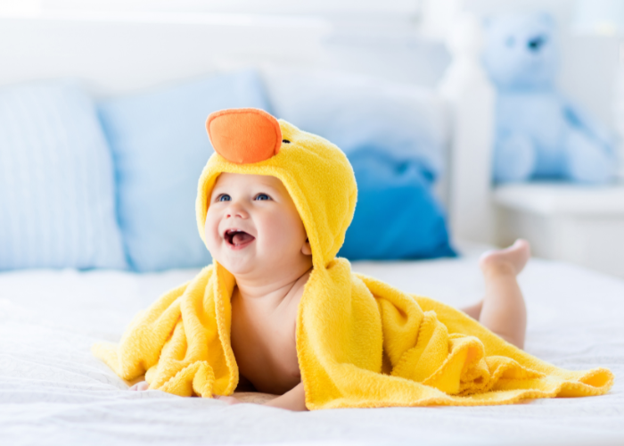 Bebê sorrindo enrolado em uma toalha de bebê amarela com capuz de patinho.